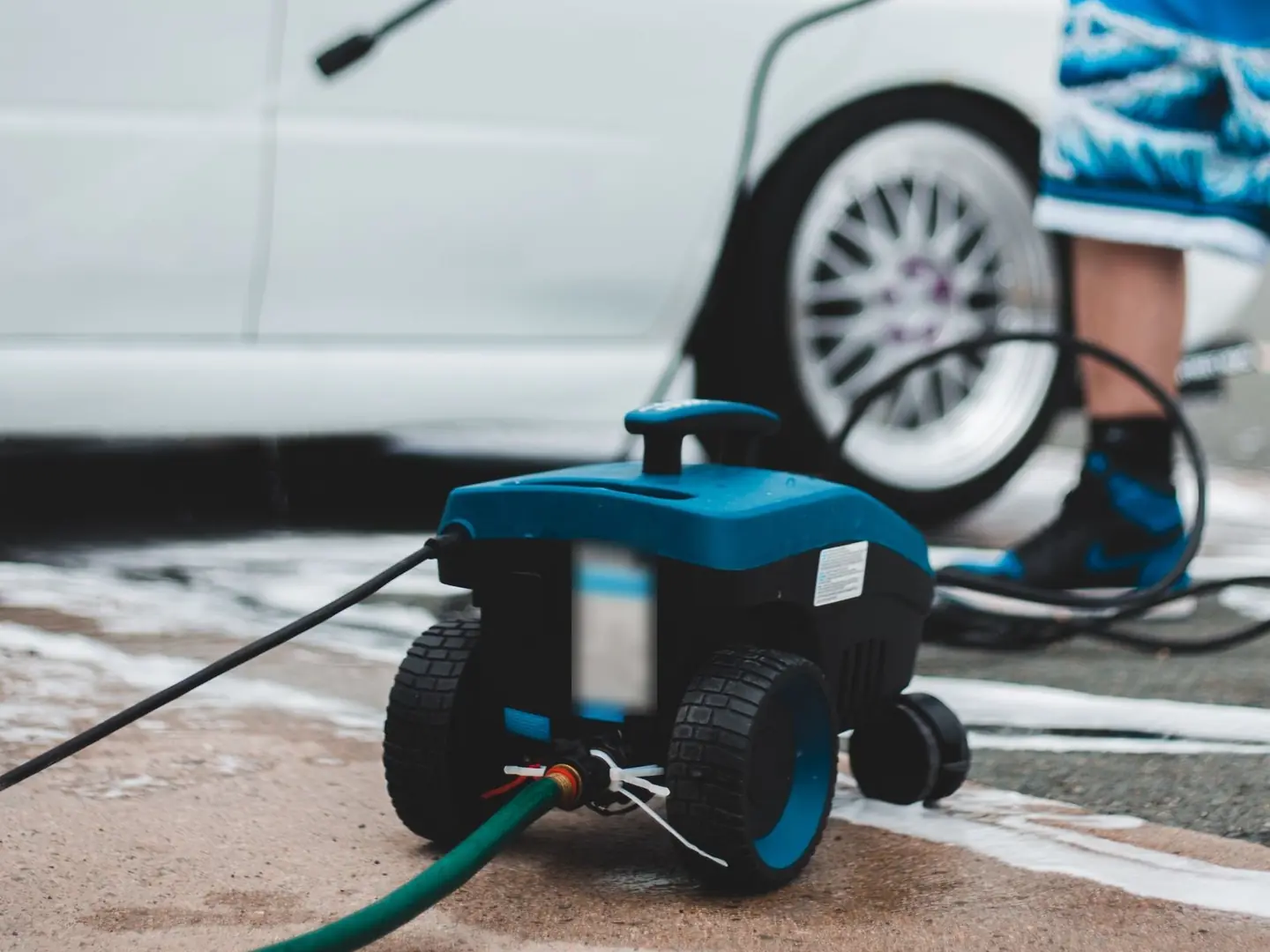 A small electric pressure washer on a wet driveway, connected to a garden hose