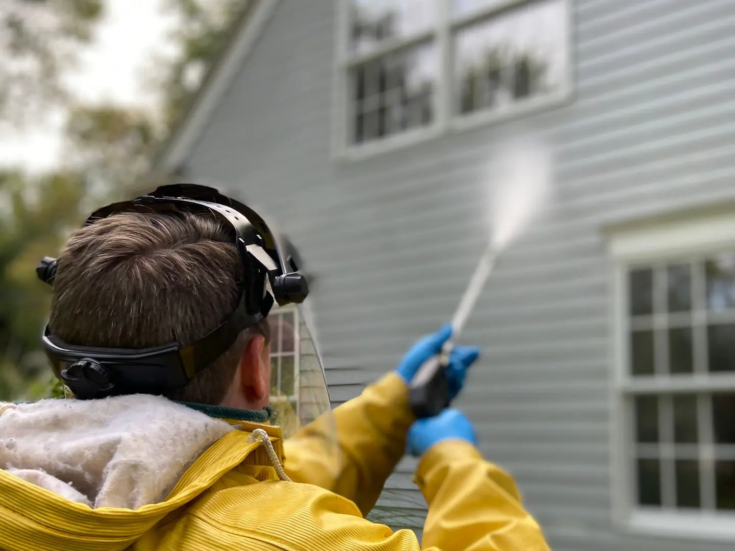 A person in a yellow rain jacket, a face shield and blue gloves pressure washing the siding of a house