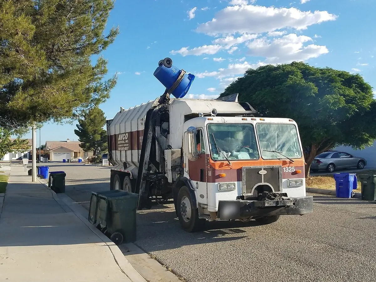An automated garbage truck’s arm lifting a blue cart on a residential street, with more carts waiting at the curb