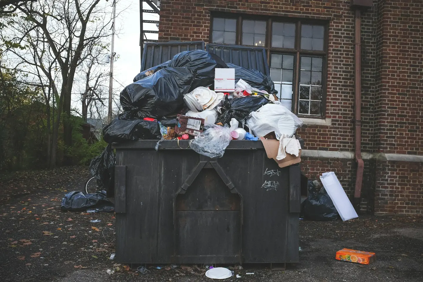 A dumpster heaped over the top with black bags and boxes beside a brick building, with litter on the ground