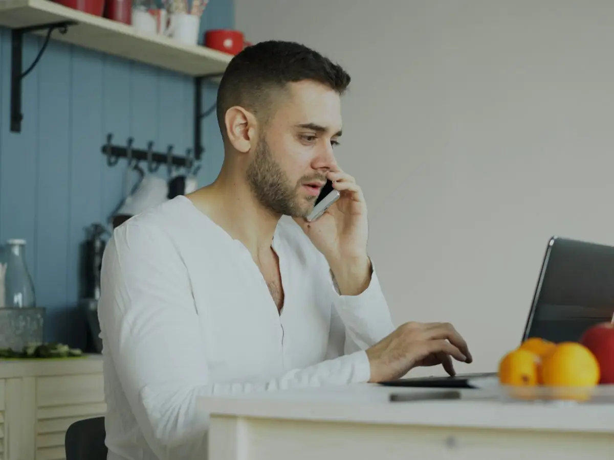 A man on a phone call while typing on a laptop at a kitchen table