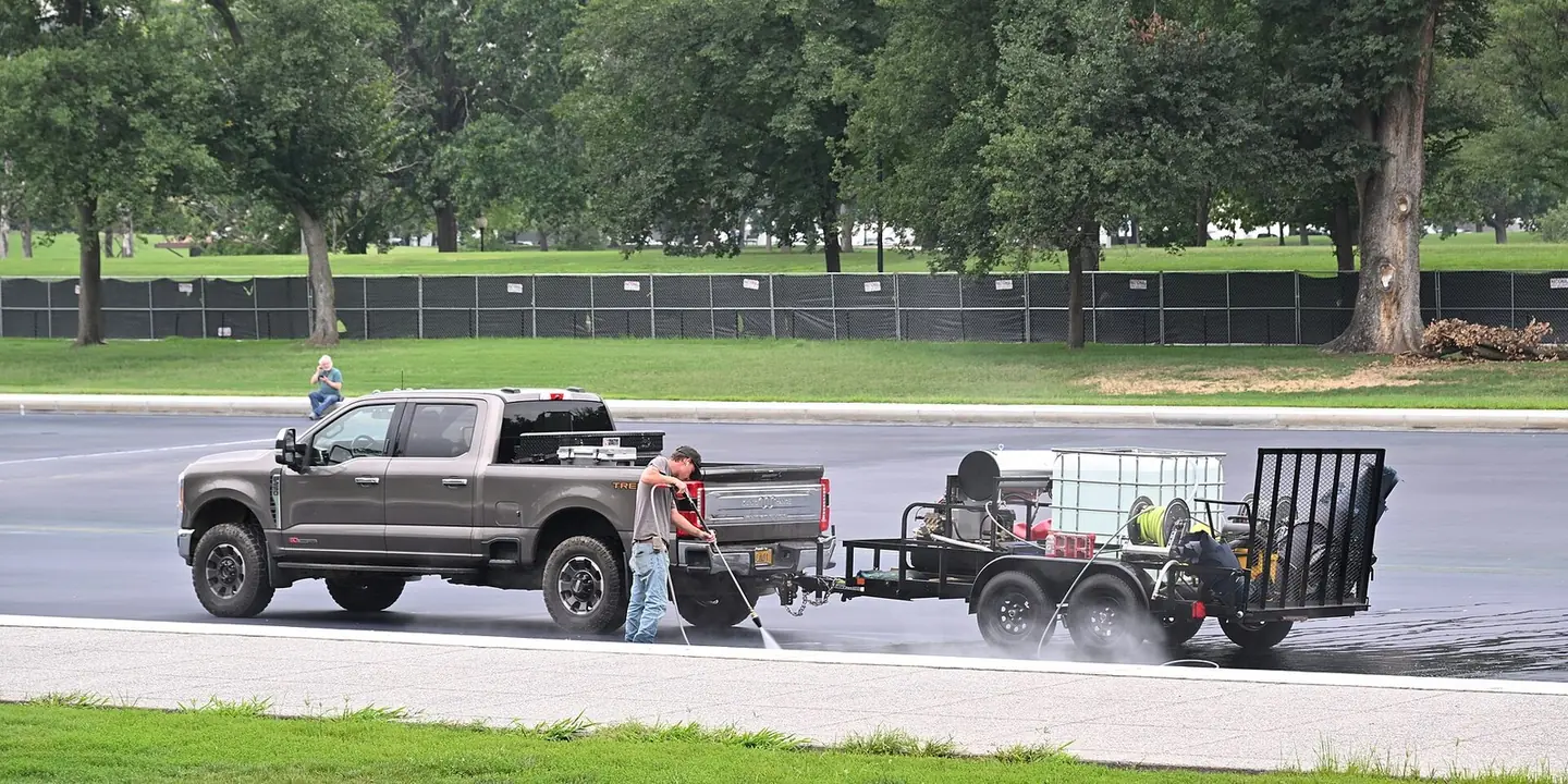 A pickup towing a flatbed trailer with a water tote, a pressure washer and a hose reel, while a worker sprays the pavement
