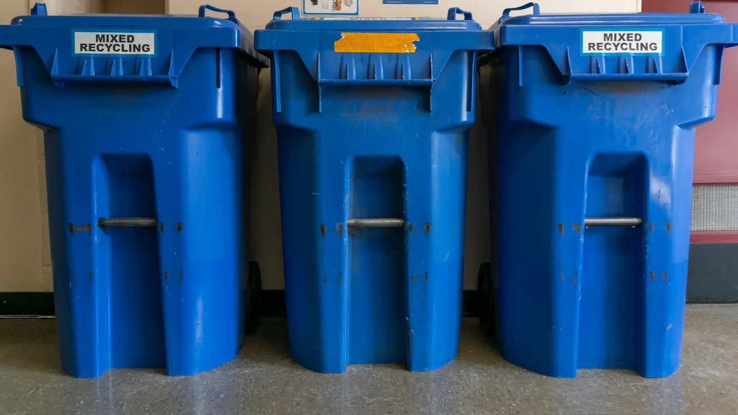 Three blue recycling carts lined up against a wall indoors