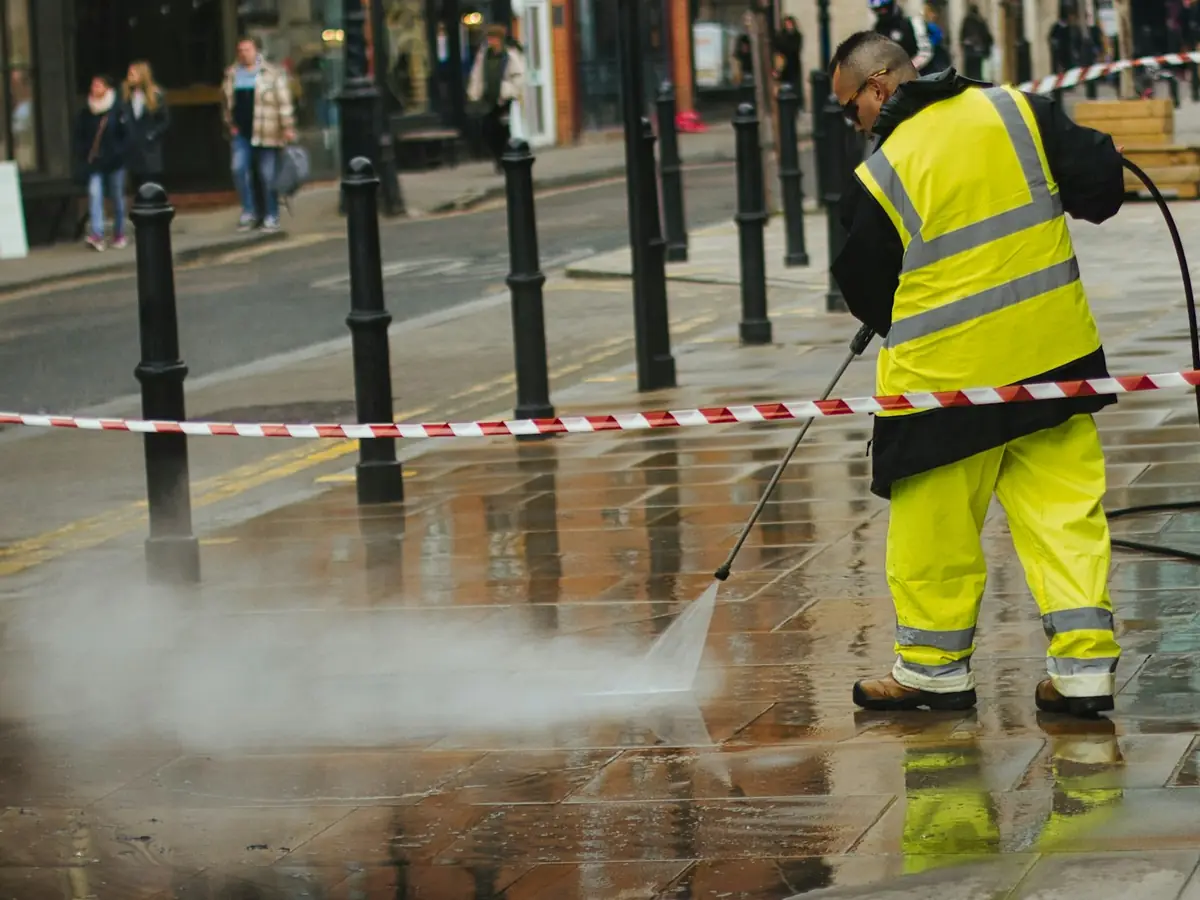 A worker in hi-vis gear pressure washes a sidewalk behind barrier tape as the wash water spreads across the pavement