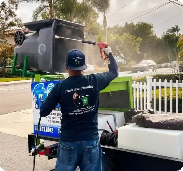 A worker washing a cart held upside down on a green tipping frame, using a wand