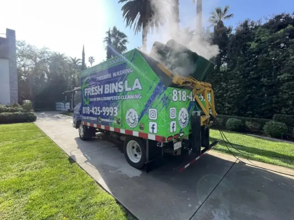 A green Fresh Bins LA truck in a driveway, its yellow lift holding a cart upside down over the wash box as steam rises